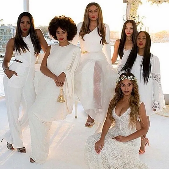 Group of six women in elegant white outfits, posing together indoors with a bright background.