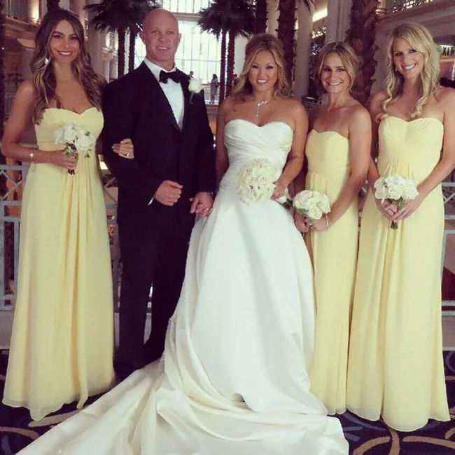 Bride and groom with bridesmaids in yellow dresses, posing indoors with palm trees in the background.