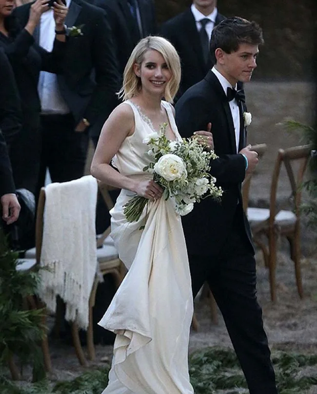 Bride in white gown holding bouquet, accompanied by a man in a tuxedo, walking down an outdoor aisle with guests.