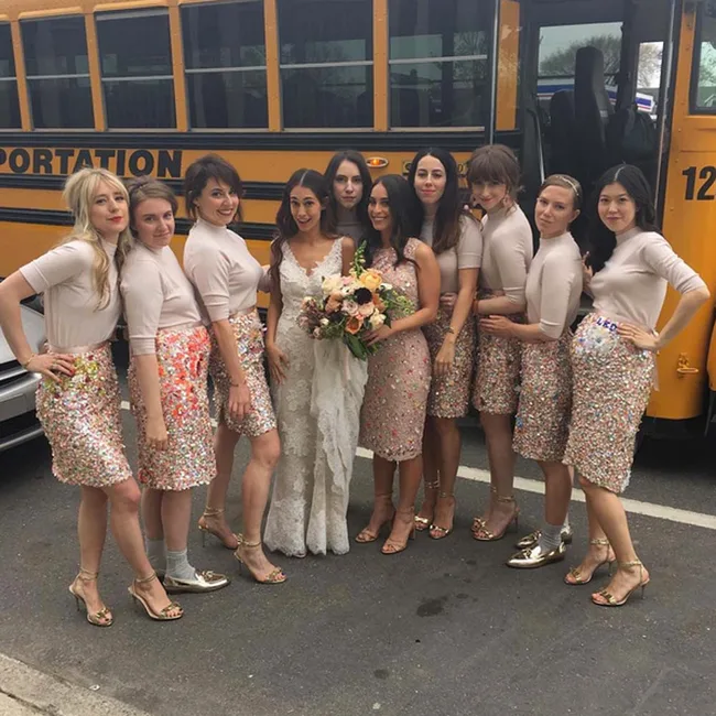 Bridal party posing in front of a school bus; bridesmaids in sequined skirts and heels, bride holding a bouquet.