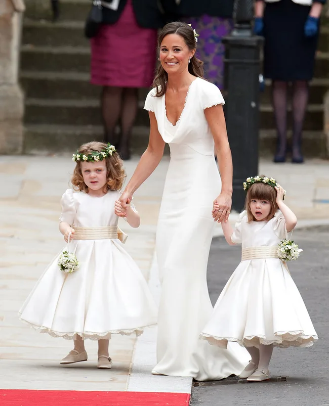A woman in a white dress walks with two young girls in flower girl dresses holding small bouquets on a stone pathway.