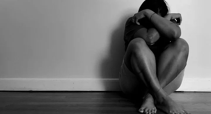 A person sits on the floor with knees drawn up, head resting on crossed arms, appearing distressed in a black-and-white photo.