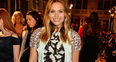 A woman with long blonde hair in a detailed dress smiling at a fashion show, surrounded by attendees.