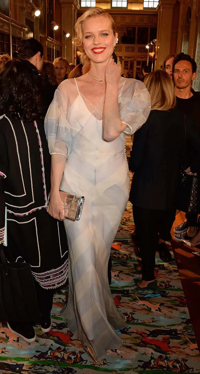 A person in a sheer white gown with a clutch, smiling at a fashion event with people in the background.