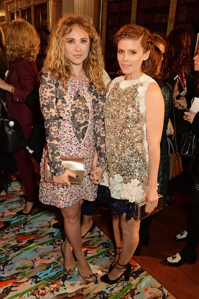 Two women in stylish dresses holding hands at an indoor event, standing on a colorful rug with people in the background.