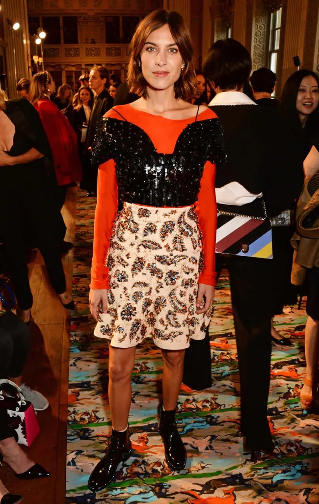 A woman in a stylish, sequined outfit stands at a fashion event, surrounded by people.