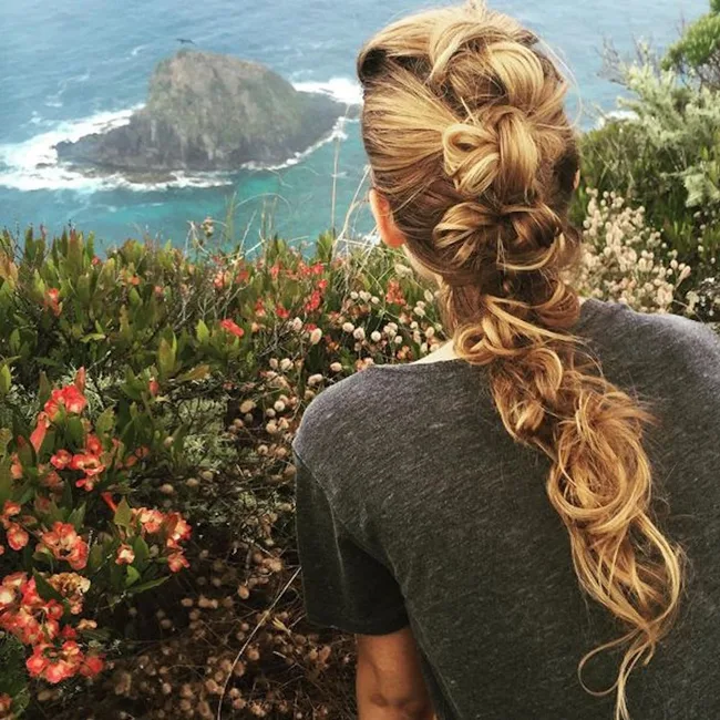 A person with intricate braided hair, sitting on a cliffside overlooking the ocean and a small rocky island.