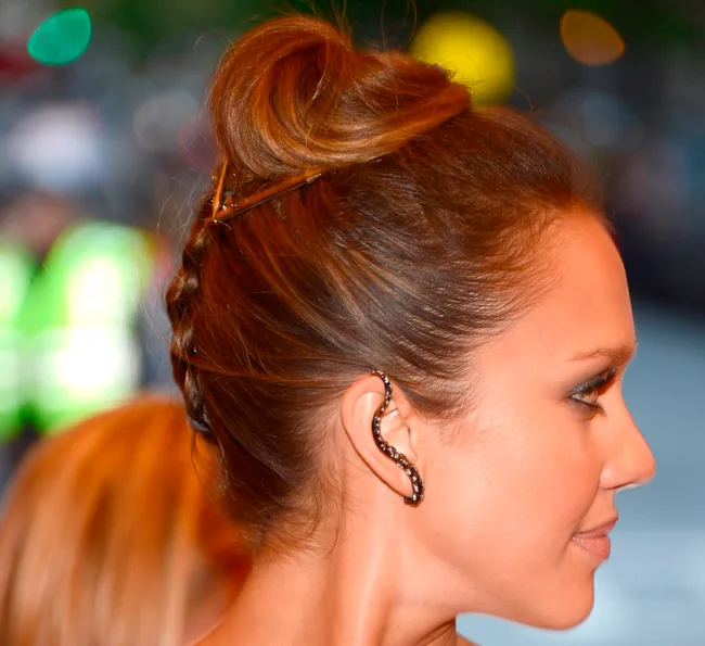 A woman with a braided updo hairstyle and a snake-shaped ear cuff, seen in profile against a blurred background.
