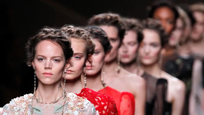 Models in elegant dresses walk the runway at a fashion show, with focus on the leading model wearing floral design.