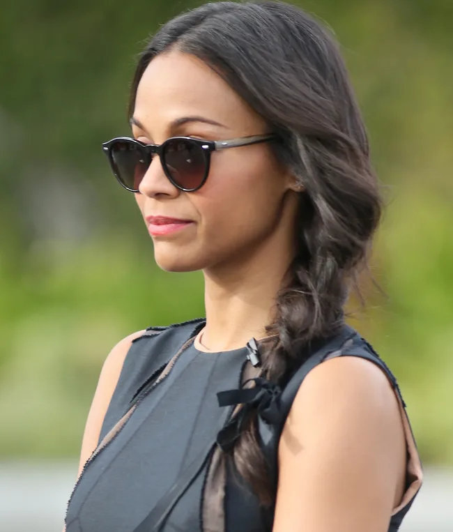 A woman with long dark hair in a braid, wearing sunglasses and a sleeveless black dress, outdoors.