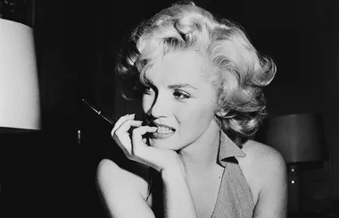 A black and white photo of a woman with curly blonde hair biting her nails thoughtfully, wearing a halter dress.