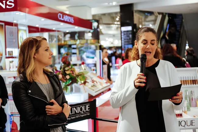 Two women speaking at a beauty event in a store, with one holding a microphone and the other listening.