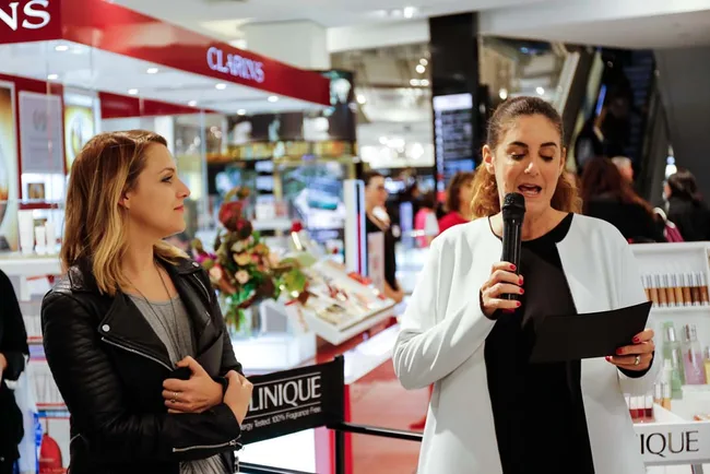 Two women speaking at a beauty event in a store, with one holding a microphone and the other listening.