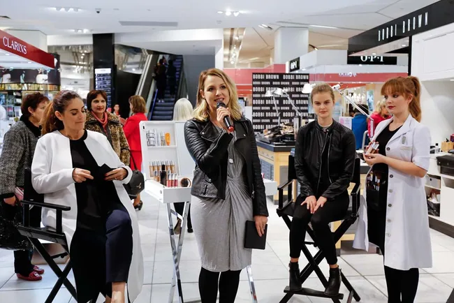 A woman speaks into a microphone at a Bobbi Brown makeup event, with others seated and standing around her.