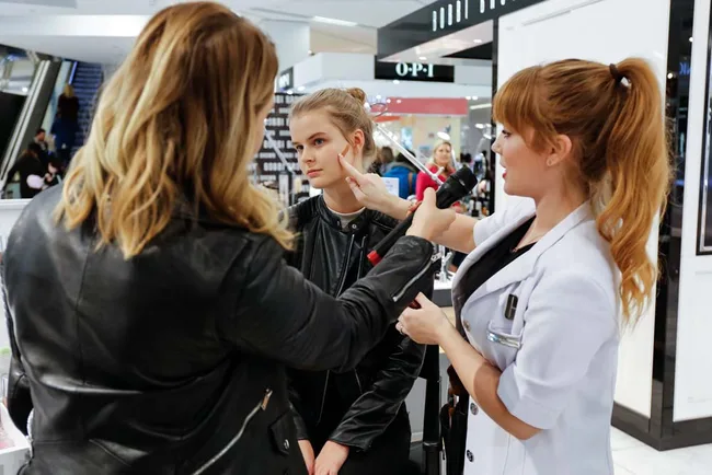 Makeup artist applying foundation on a seated woman's face while another woman observes in a mall setting.