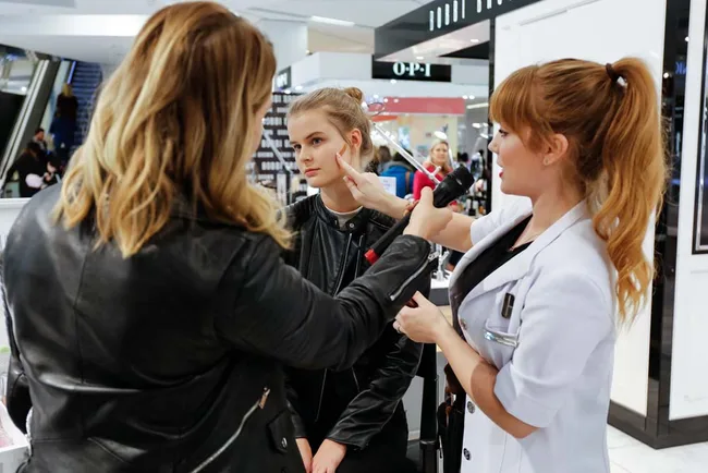 Makeup artist applying foundation on a seated woman's face while another woman observes in a mall setting.
