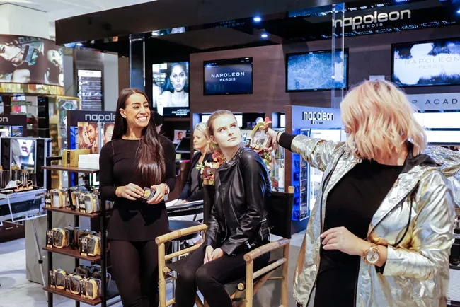 Makeup demonstration at Napoleon Perdis store with two women involved, vibrant shelves in the background.