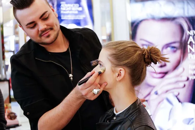 A makeup artist applying foundation on a woman's face near a beauty product advertisement.