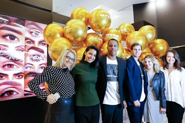 Group of six people smiling, standing in front of golden balloons and an eye-themed display in a well-lit area.