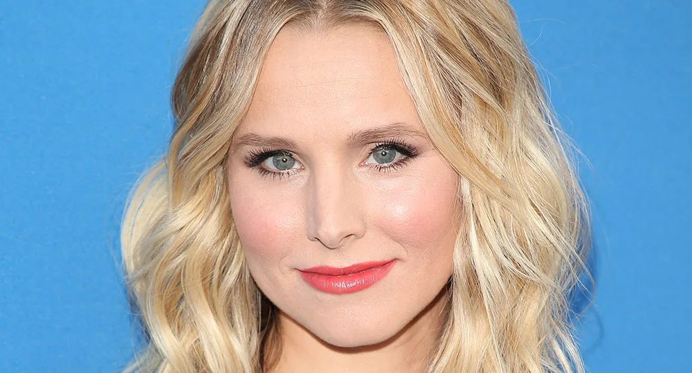 Blonde woman with wavy hair and a blue background, wearing subtle makeup and a gentle smile.