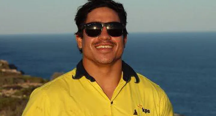 Man wearing a yellow shirt and sunglasses, smiling with the ocean in the background.