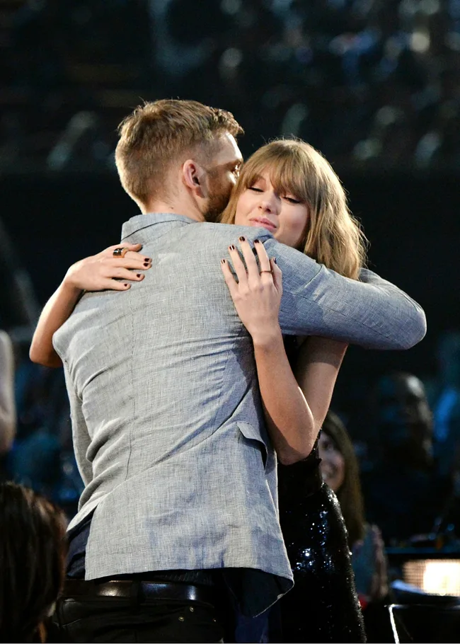 A man in a gray suit hugs a woman in a black dress at an event, with blurred audience in the background.