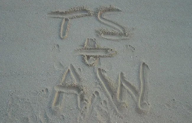 Sand letters reading "T S + A W" with small footprints around on a beach.