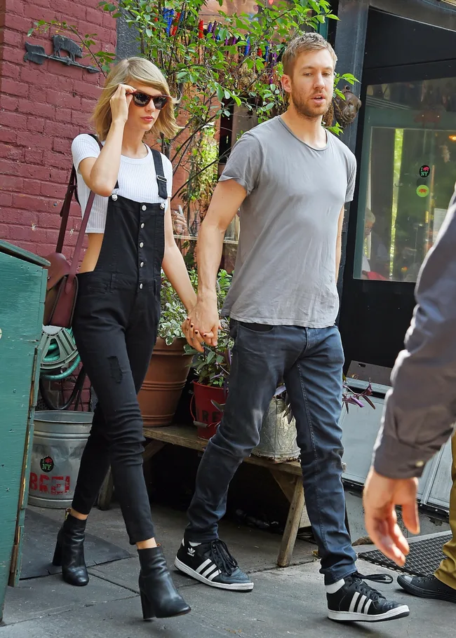 A person in black overalls and sunglasses holds hands with another in a gray shirt on a street near plants.