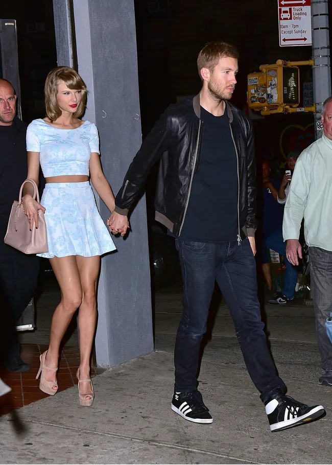 Two people holding hands on a sidewalk at night, with one wearing a light blue outfit and the other in a leather jacket.