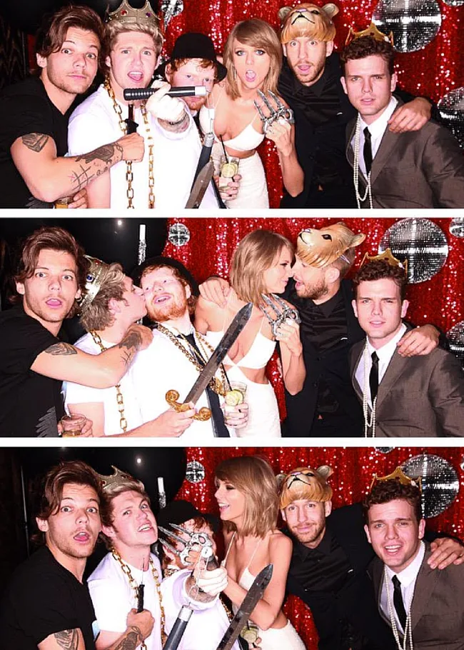 Group of people in a fun photo booth, with props like a crown, sword, bear mask, against a red glittery backdrop.