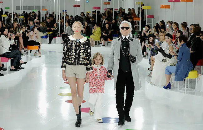 "Fashion show with two adults and a child walking down the runway, surrounded by seated audience members."
