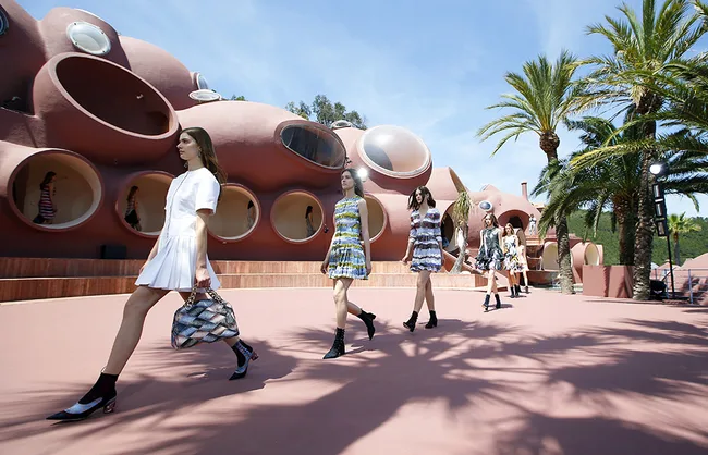 Models walk in colorful dresses at a fashion show outside a futuristic, bubble-shaped building.