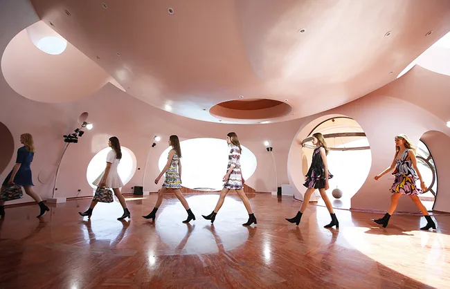 Models walking in a line wearing colorful dresses and boots inside a spacious room with circular walls.