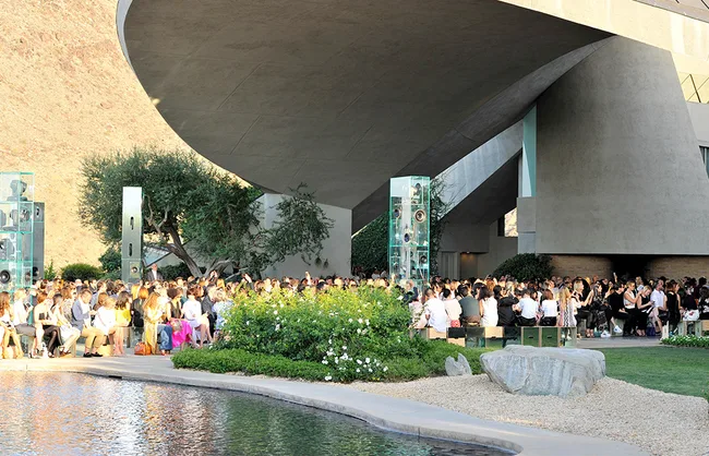 Outdoor gathering at modern architectural venue with a large crowd seated near a pond.