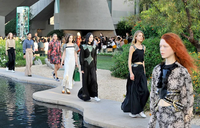 Models walking on a path near a pond during an outdoor fashion show.