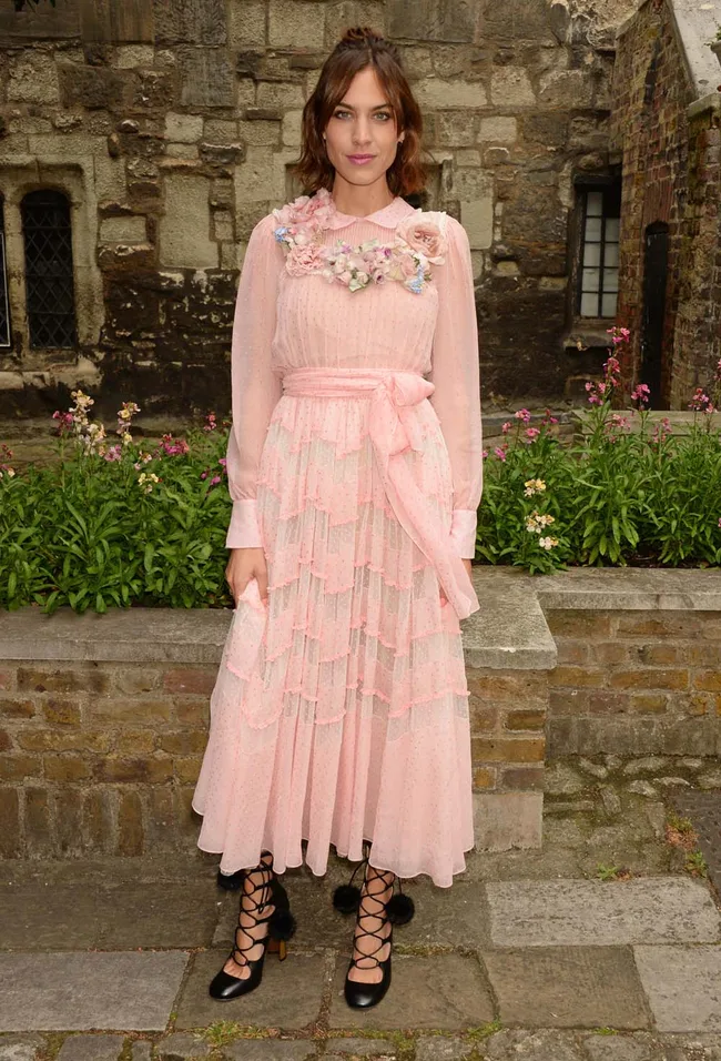 A woman in a pink floral dress with lace-up shoes stands outdoors against a stone wall and colorful flowers.
