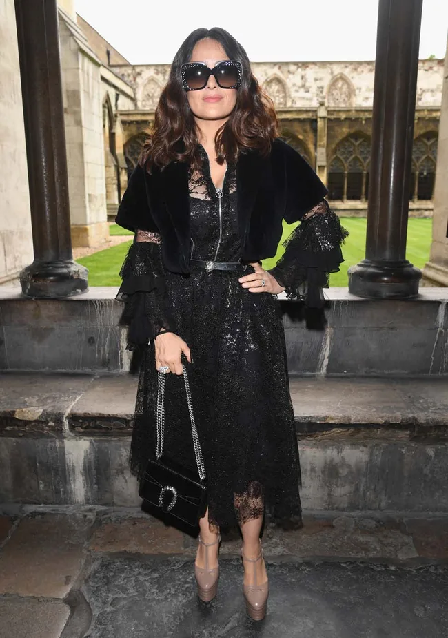 A woman in a black lace dress with sunglasses stands in a stone courtyard.