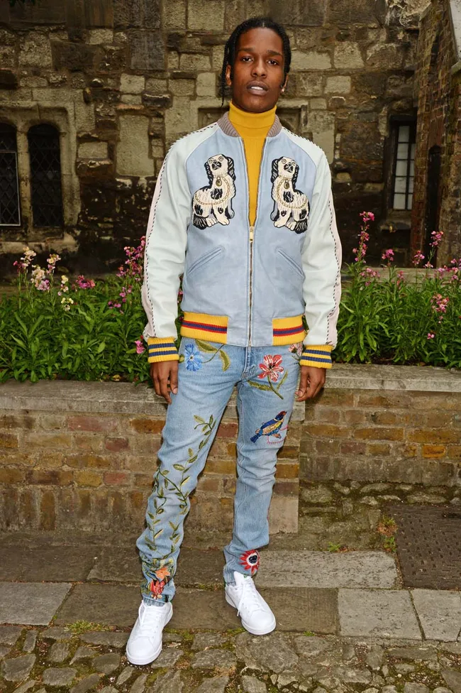 Person in a stylish jacket with dog prints and embroidered jeans, standing by a stone wall and flowers.