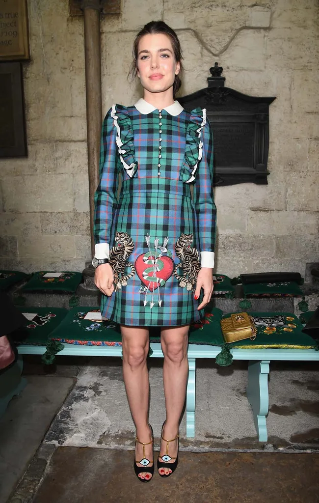 Person in a plaid dress with embroidery, standing indoors beside stone walls and cushioned pews.