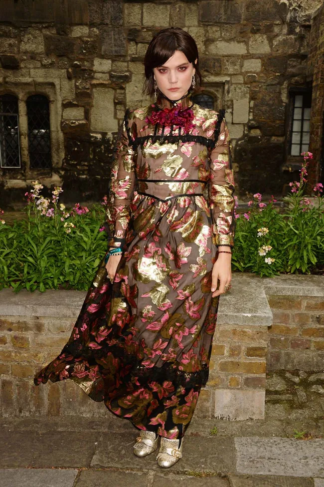 Woman in a floral gold and pink dress stands in front of a stone building with flowers.