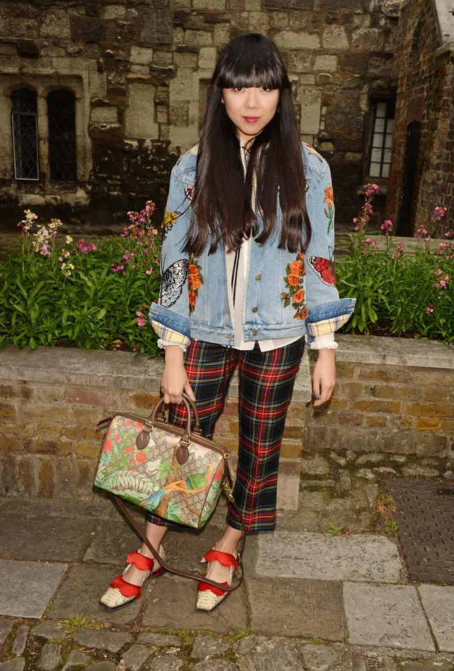 Young woman in embroidered denim jacket and plaid pants, holding a floral-patterned bag, stands in a garden setting.