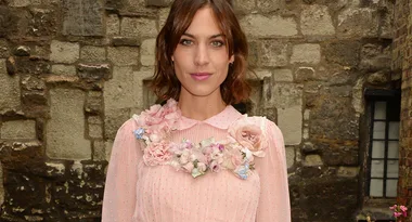 A woman in a pink dress with floral embellishments stands in front of a stone wall.