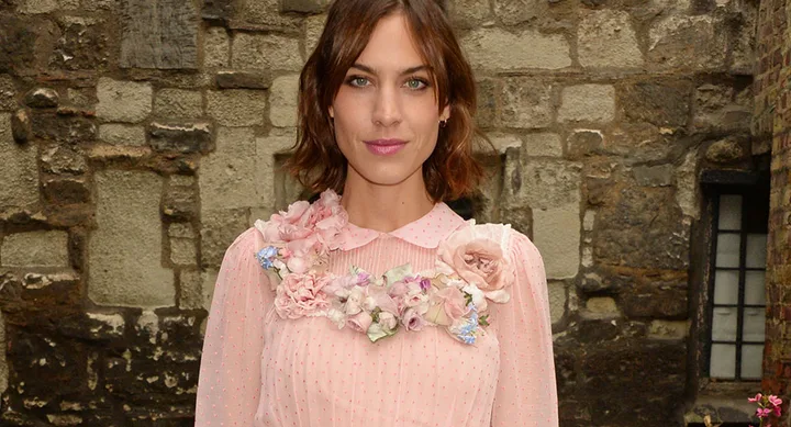 A woman in a pink dress with floral embellishments stands in front of a stone wall.