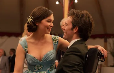 Young woman in a blue dress sits on man's lap, both smiling at each other warmly, in a festive setting.