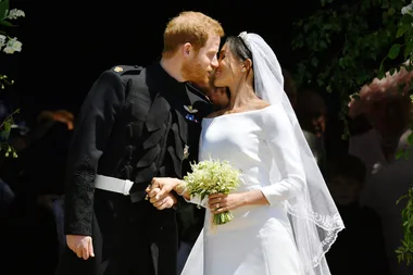 A bride and groom in formal attire share a kiss outside a venue, surrounded by greenery and holding a bouquet.