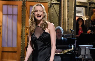 ```plaintext
A woman in a black dress smiles on a stage, with a band visible in the background.
```