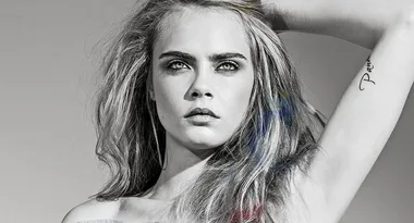 Grayscale portrait of a woman with long hair, looking into the distance, showing a tattoo on her arm.