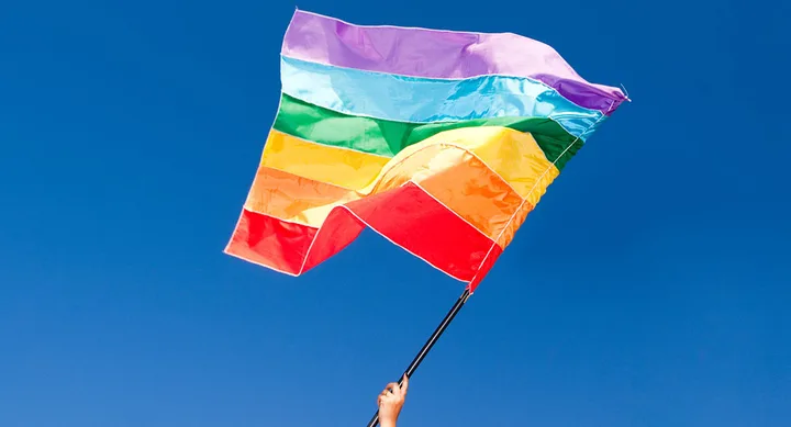 A hand holds a waving rainbow pride flag against a clear blue sky.