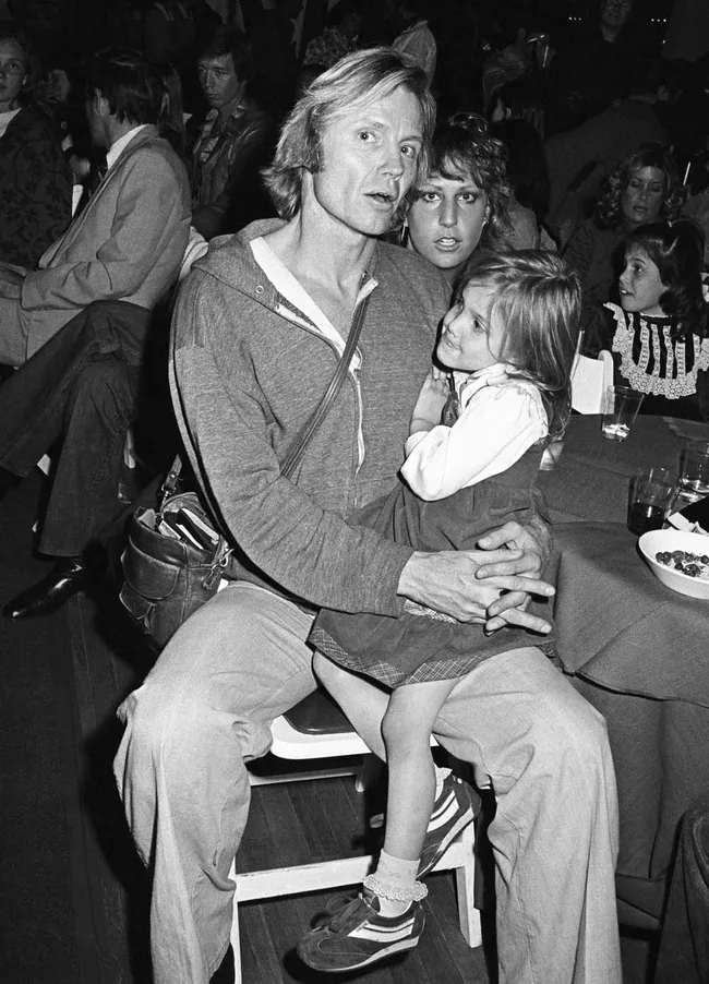 Man in casual attire holds a young girl on lap at a crowded event with people seated around them.