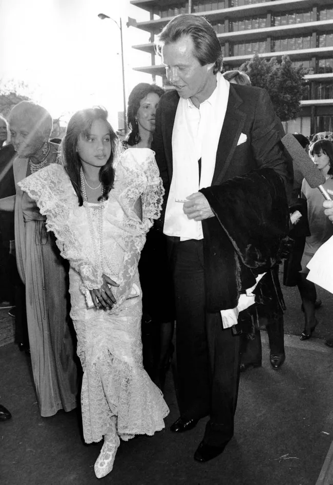 A young girl in a lace dress and a man in a suit stand together at an outdoor event, surrounded by a crowd.